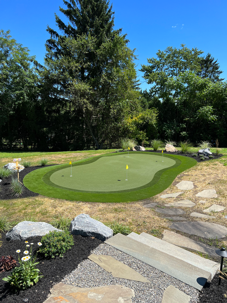 A backyard putting green with two holes, surrounded by landscaping, rocks, and a stone pathway in a sunny, grassy yard.
