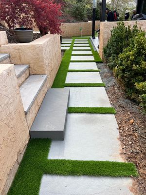 A stone walkway with square concrete pavers separated by strips of artificial grass, leading past a stone retaining wall.