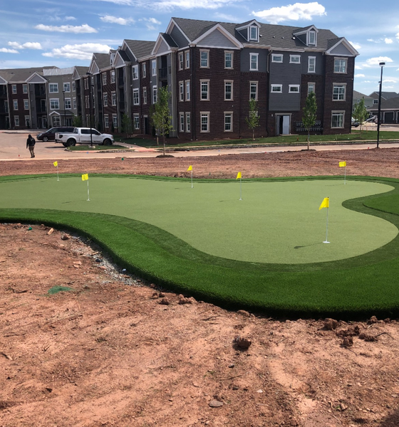 An artificial putting green with yellow flags sits in front of a multi-story apartment building under a blue, cloudy sky.