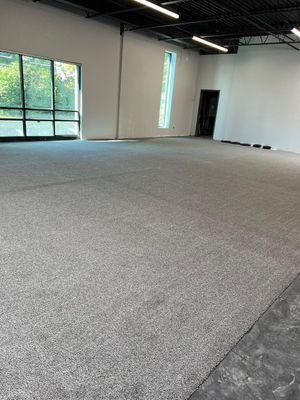 A spacious, empty room with grey carpet, white walls, tall windows, and a doorway under a black ceiling with lighting.