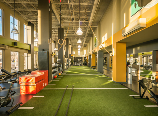 Wide-angle shot of a gym with a green turf strip, black columns, bright orange plyo boxes, and hanging gymnastic rings.