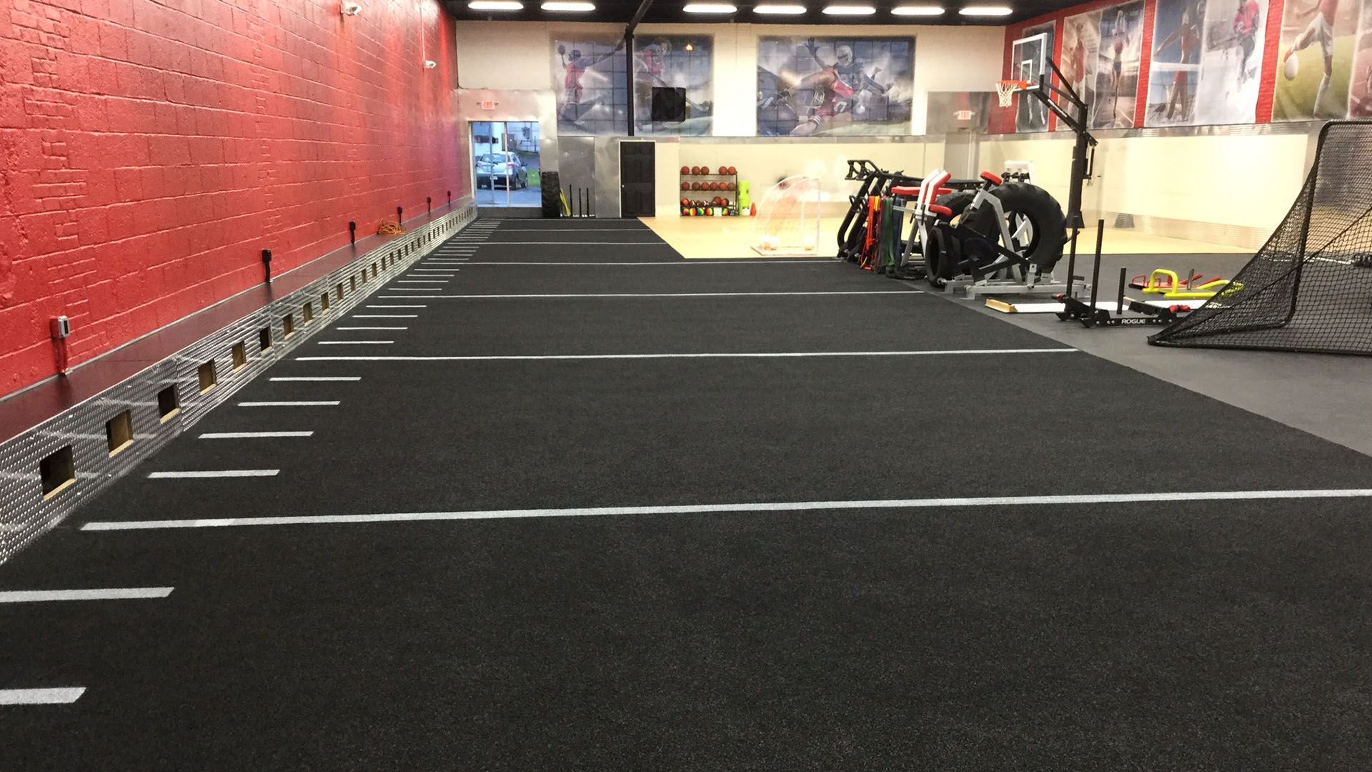 An indoor sports facility with a long black turf track featuring white yard lines along a red wall.