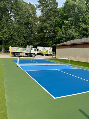 A blue and light blue pickleball court with a net, set against a backdrop of trees, a building, and construction trucks.