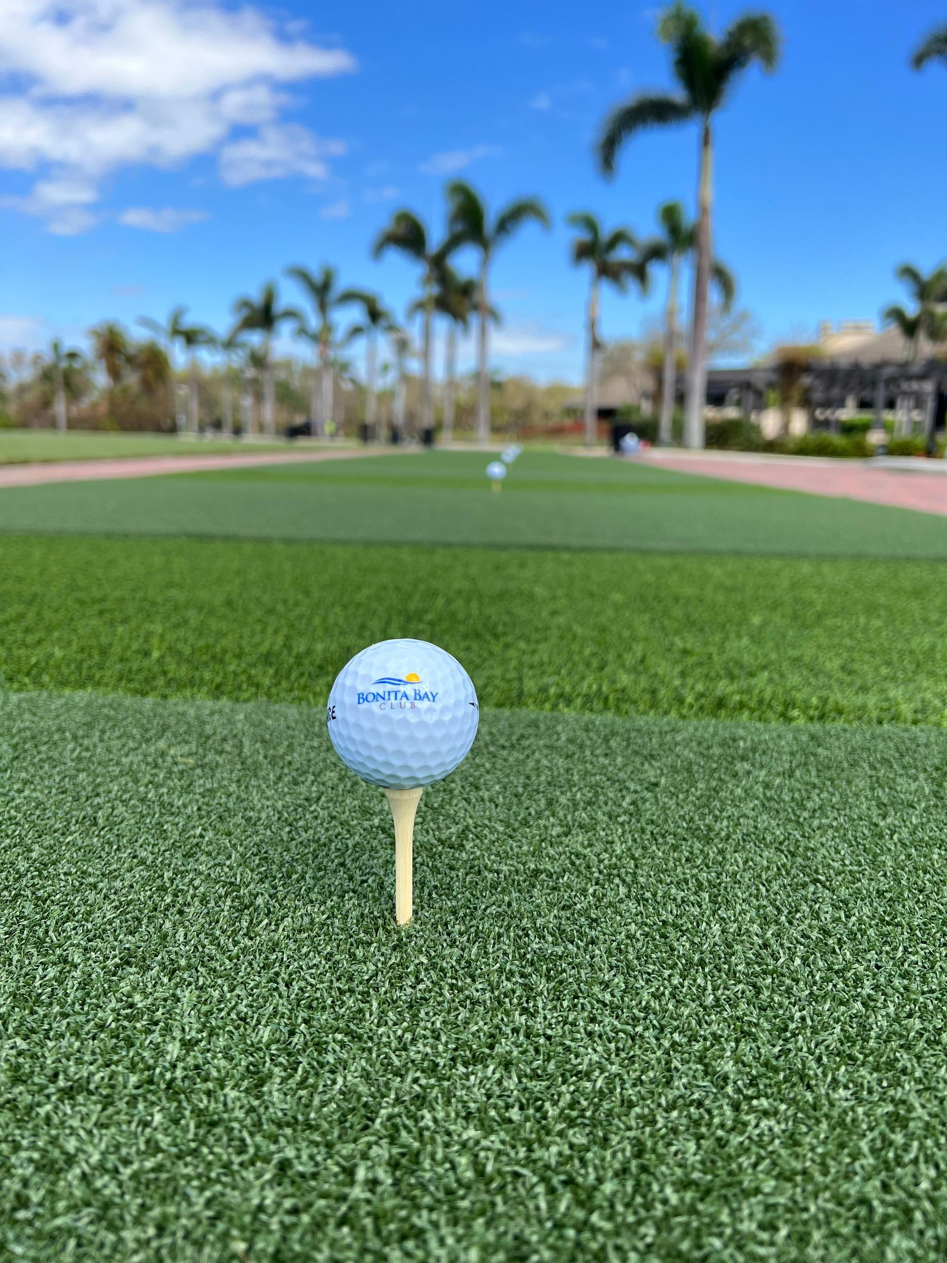 A light blue golf ball on a white tee sits in the foreground of a manicured putting green under a bright, sunny sky.