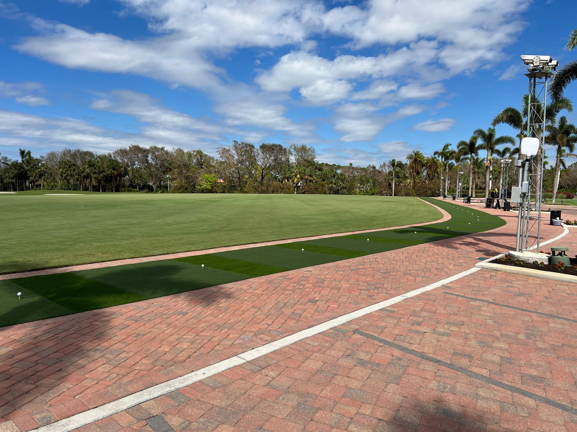 A view of a golf practice facility with a paved brick walkway leading to artificial turf hitting stations and a lawn.