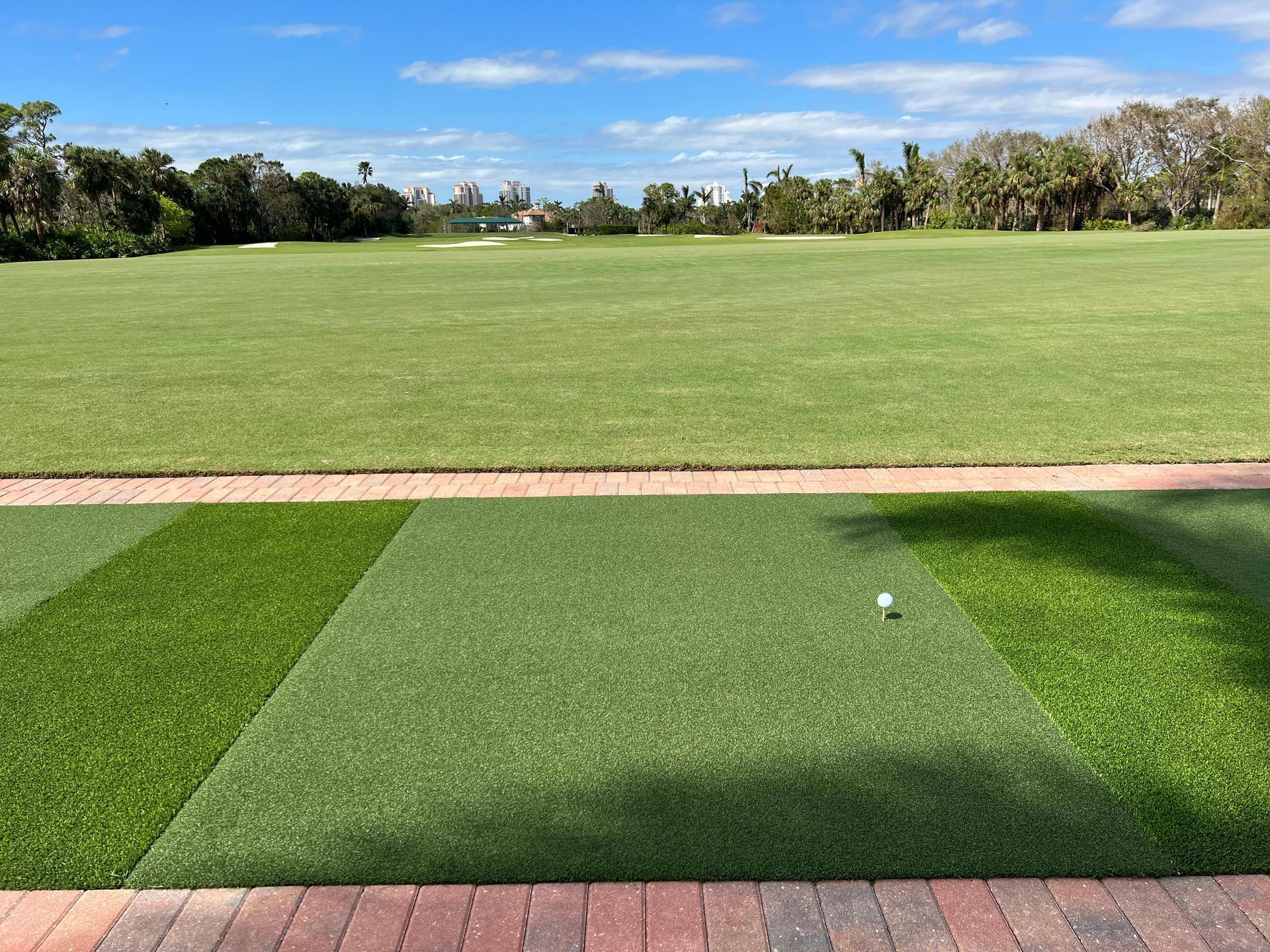 A golf ball sits on an artificial turf tee area, looking out over a wide, grassy driving range under a bright blue sky.