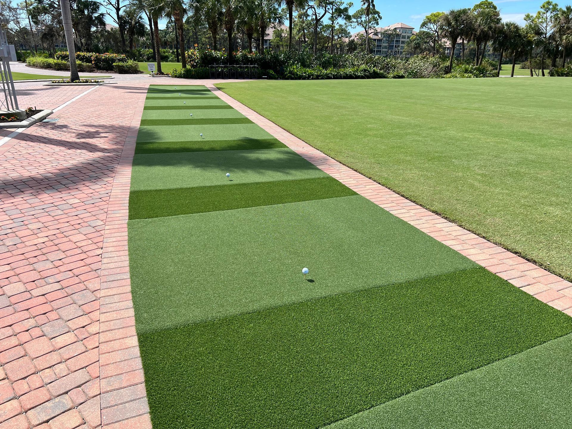 A golf driving range mat with a single white golf ball, bordered by a red brick walkway and a large grassy field.