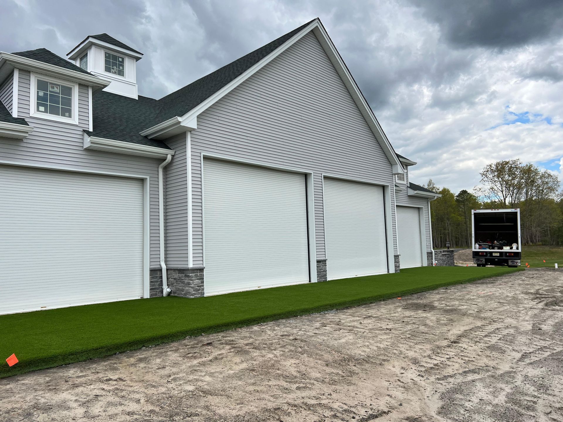 A light gray garage building with white roll-down doors, a shingled roof, and new green turf against a dirt yard.