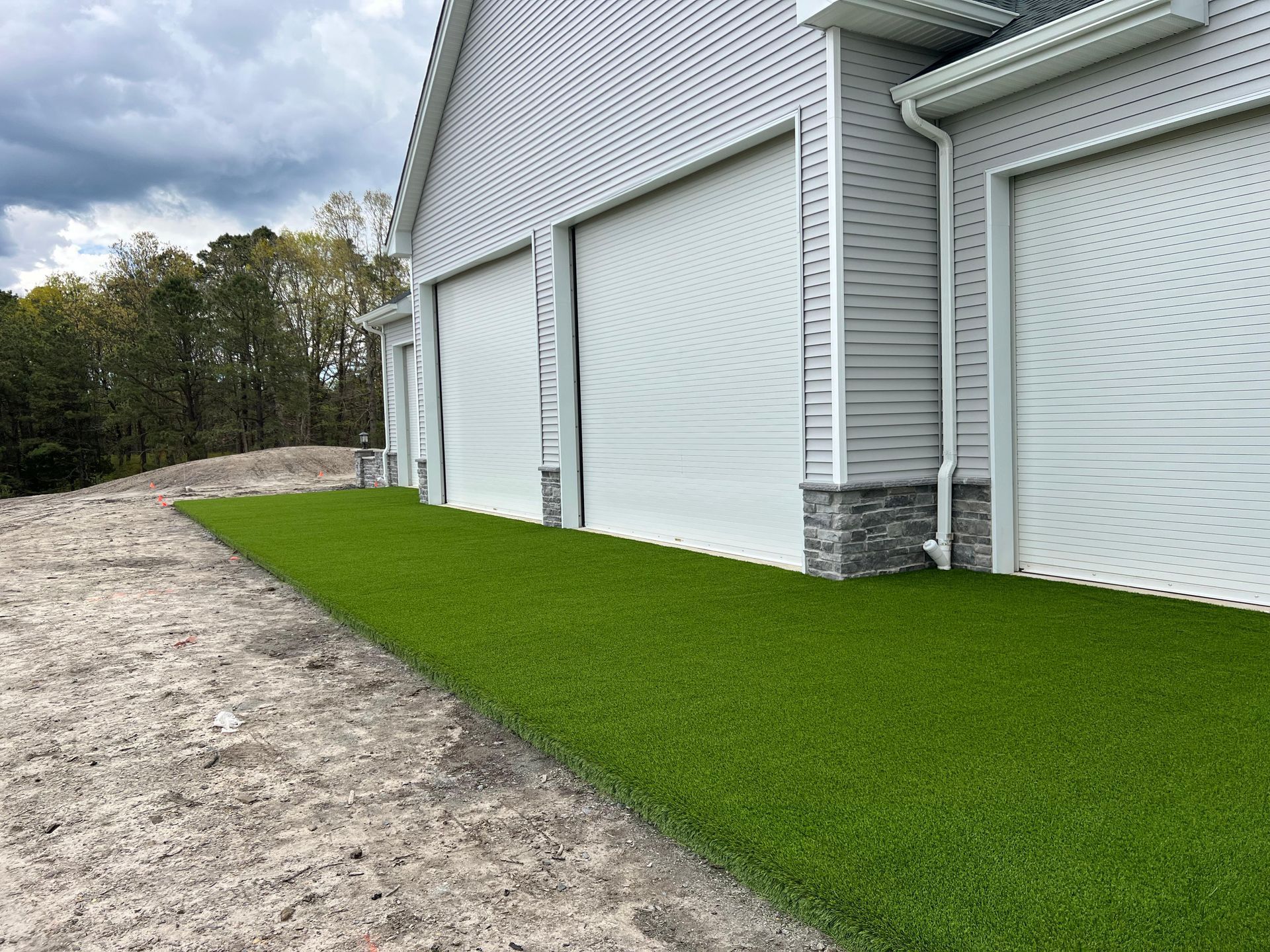 A bright green artificial lawn sits next to a light-colored garage with three closed white roll-up doors.