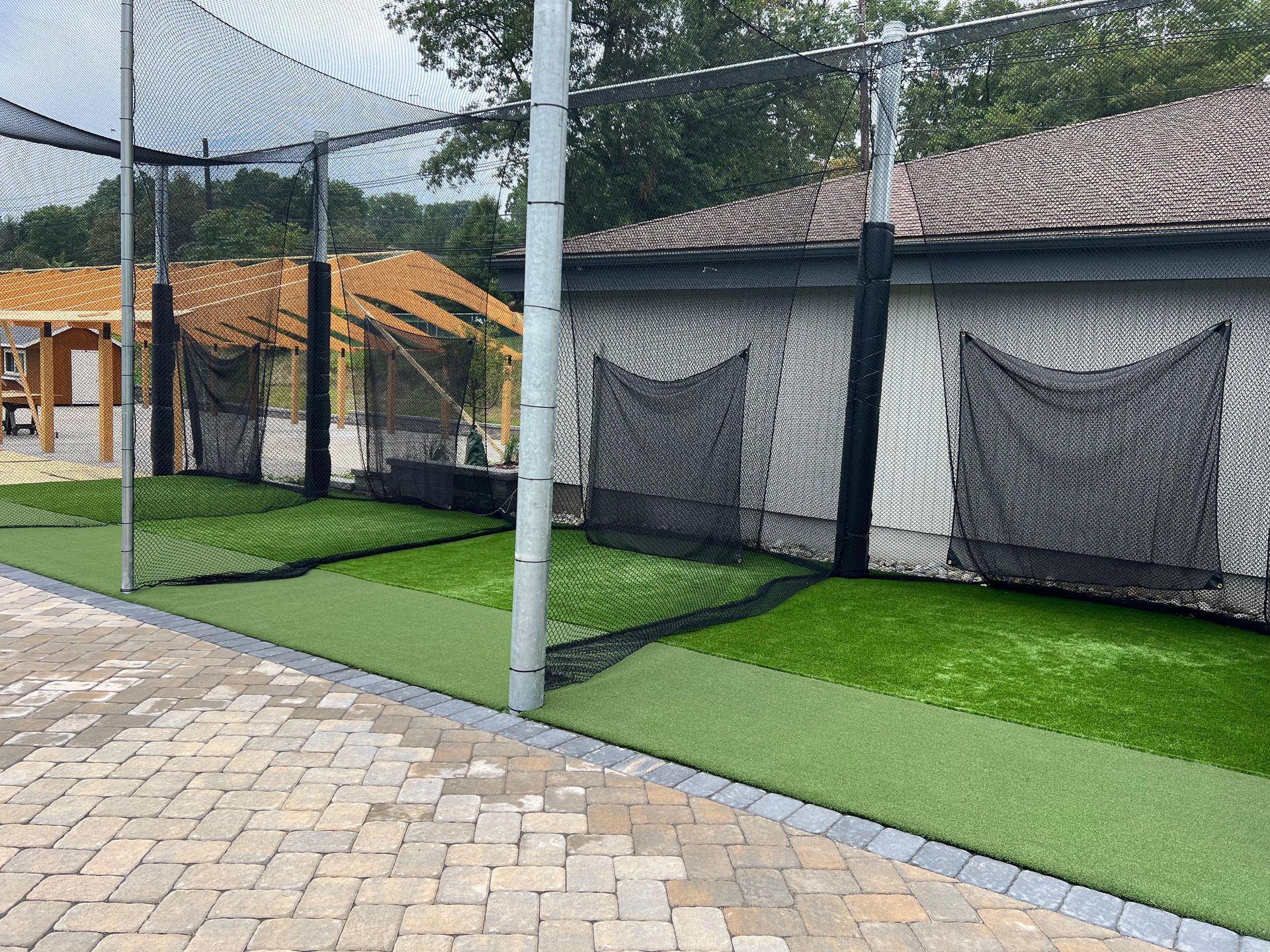 Outdoor batting cage setup with green artificial turf, black netting, and padded posts next to a building.