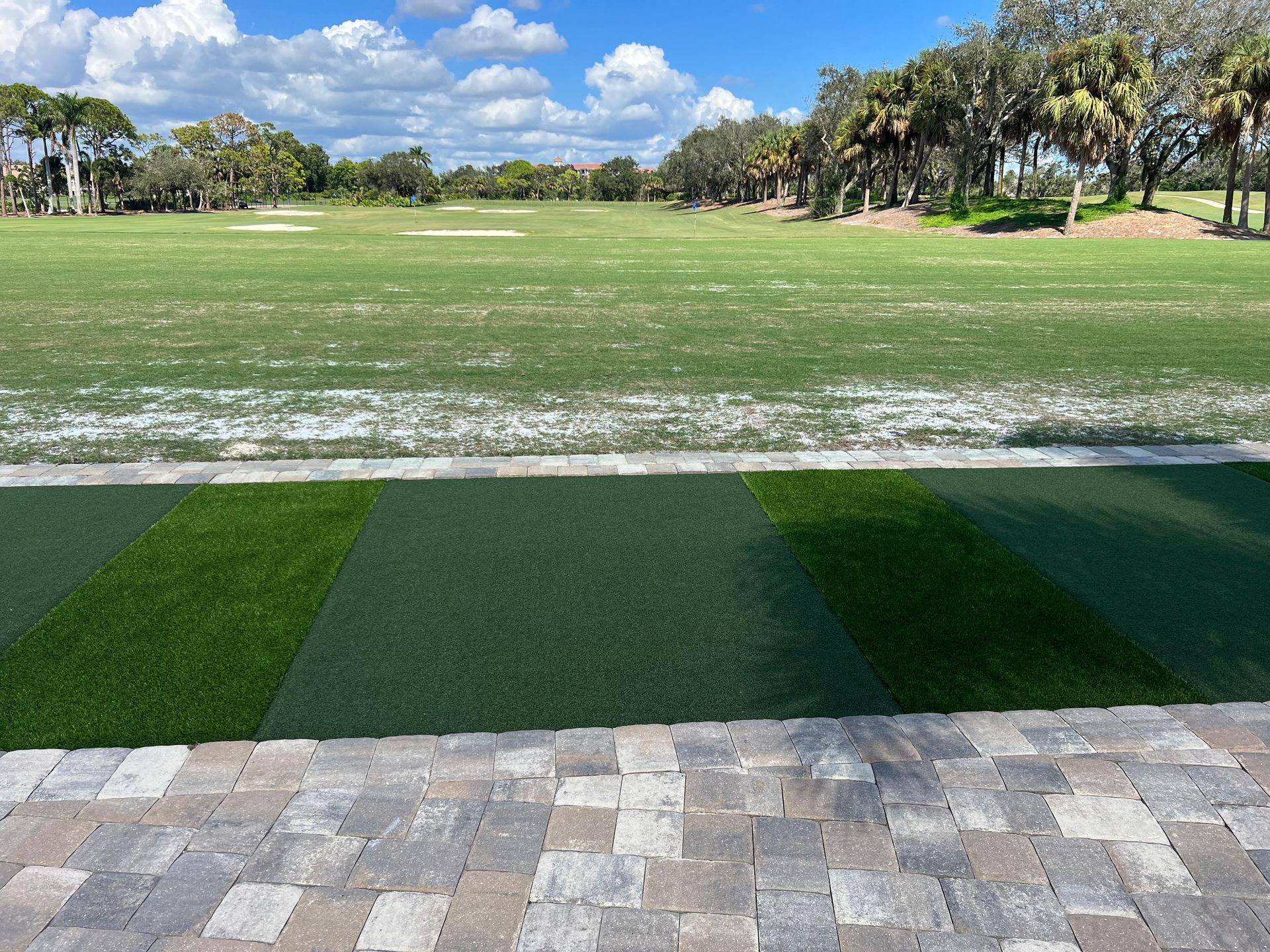 A view from a paved patio looking out onto a large, open green golf driving range under a partly cloudy blue sky.