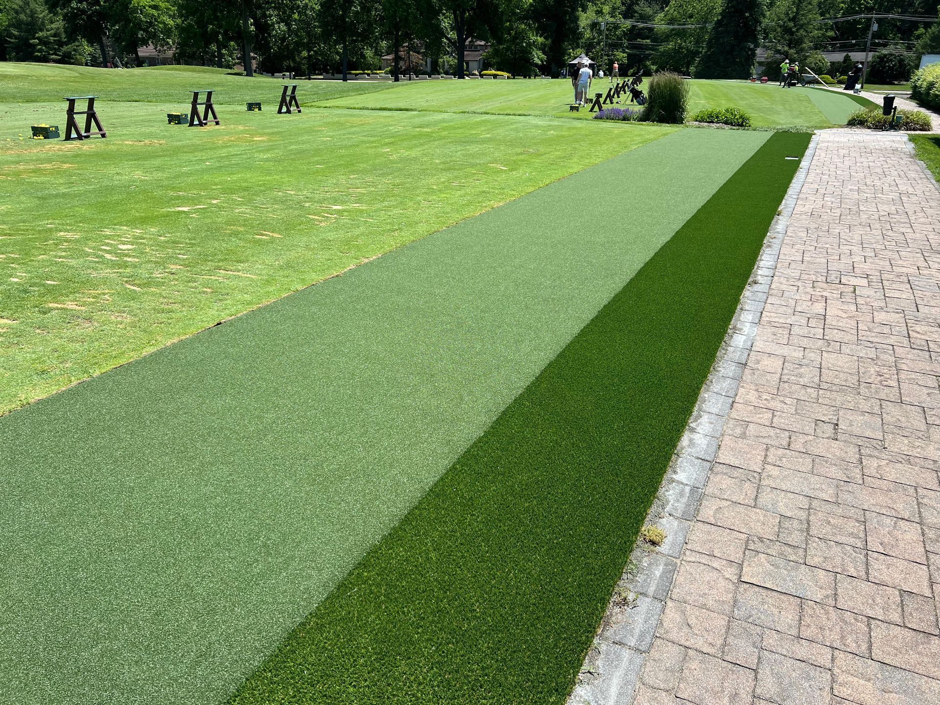 A path transitions from a brick walkway to two strips of different artificial turfs, leading toward a grassy golf range.