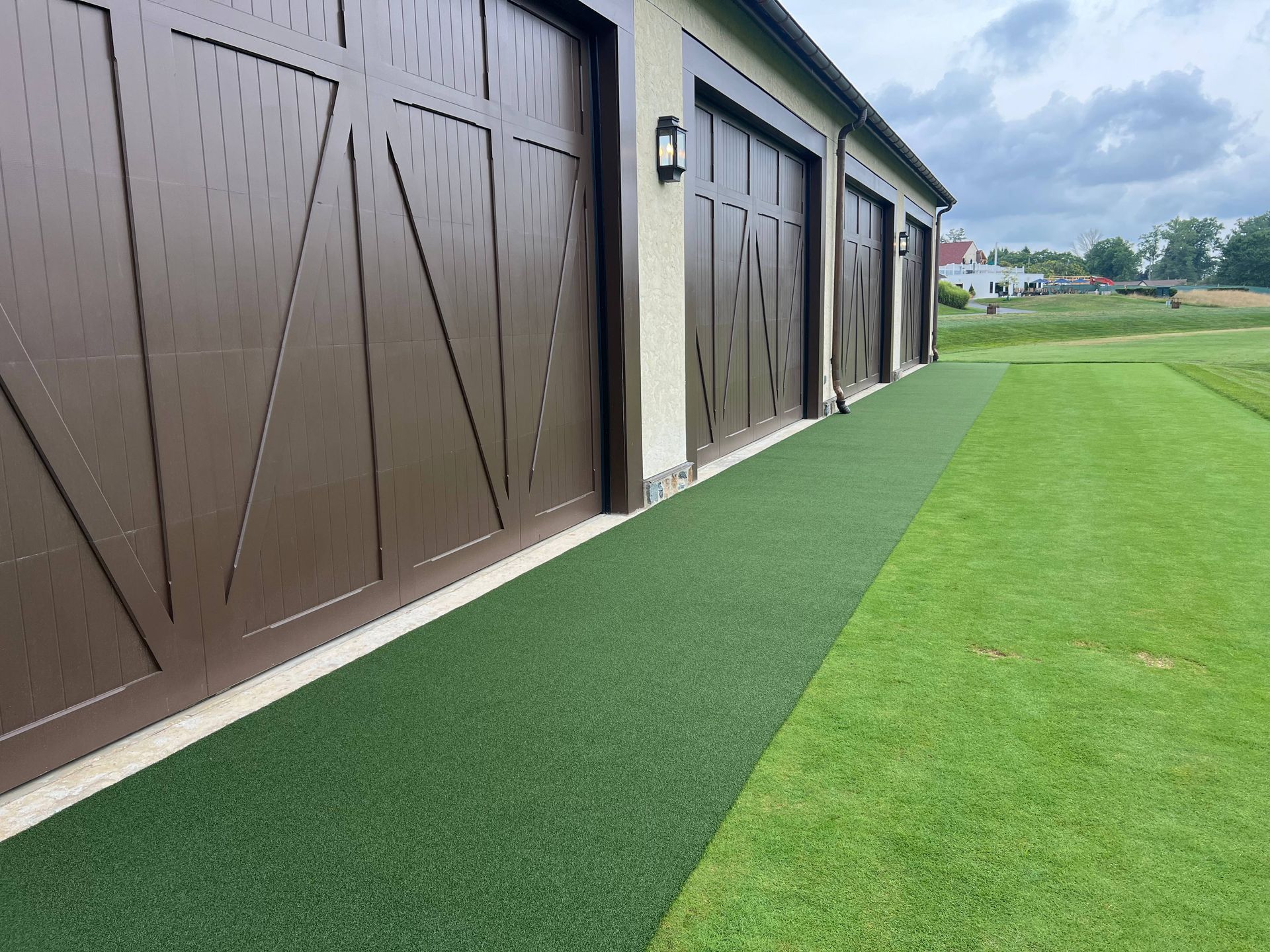 A strip of artificial turf runs along the side of a building with multiple brown garage doors, bordering a grassy lawn.