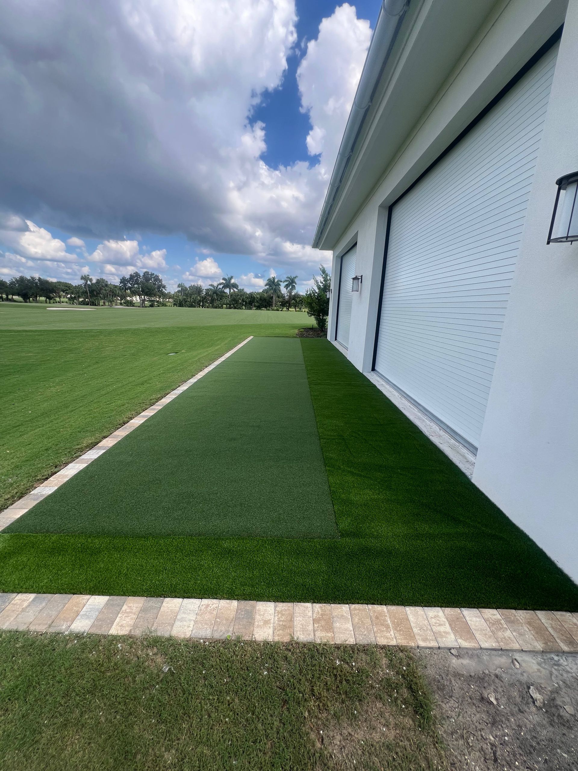 A strip of artificial turf runs along the white wall of a building, bordered by stone pavers next to a grassy lawn.