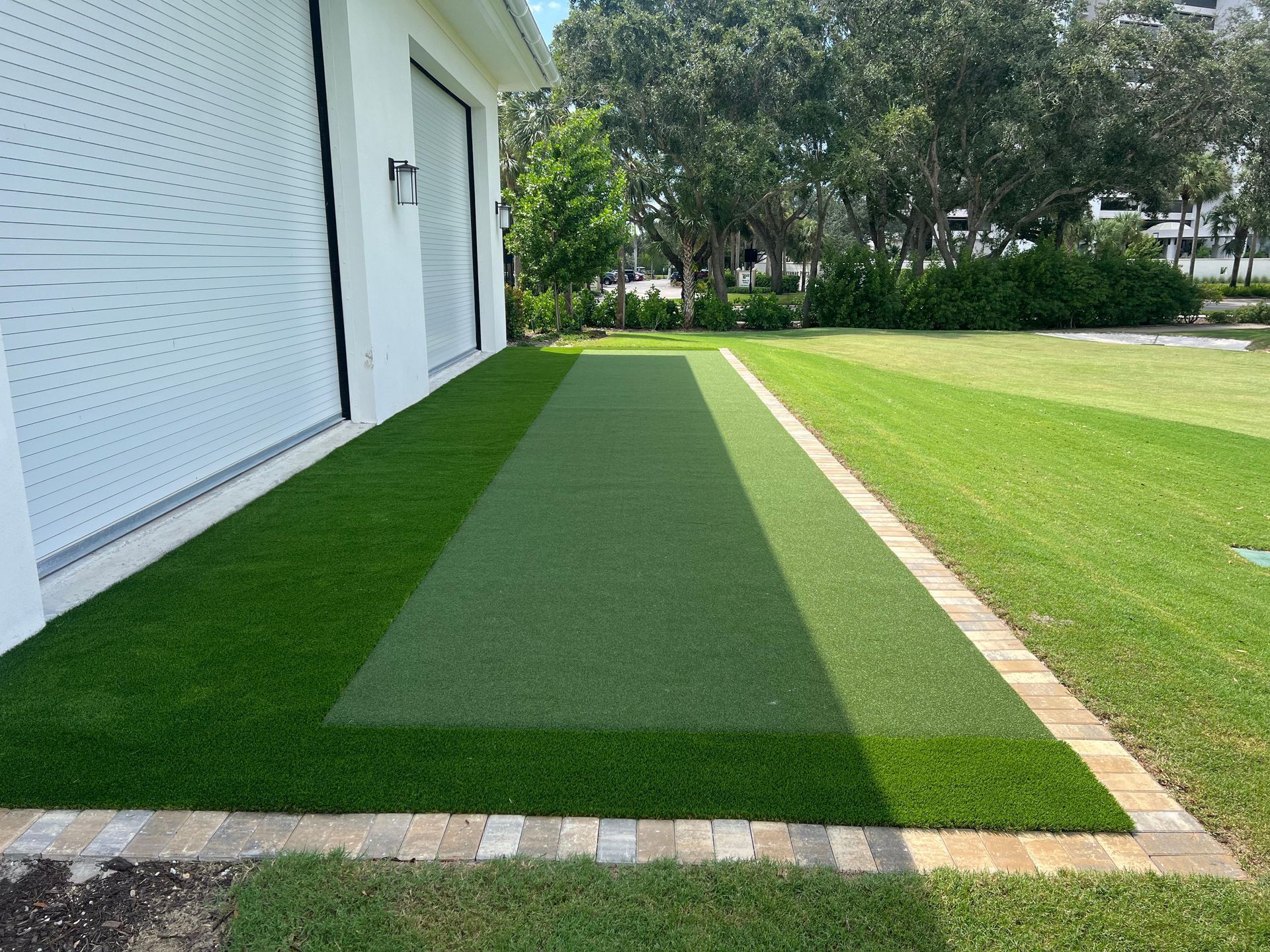 A strip of artificial turf bordered by pavers next to a white house wall, with natural grass and trees in the background.