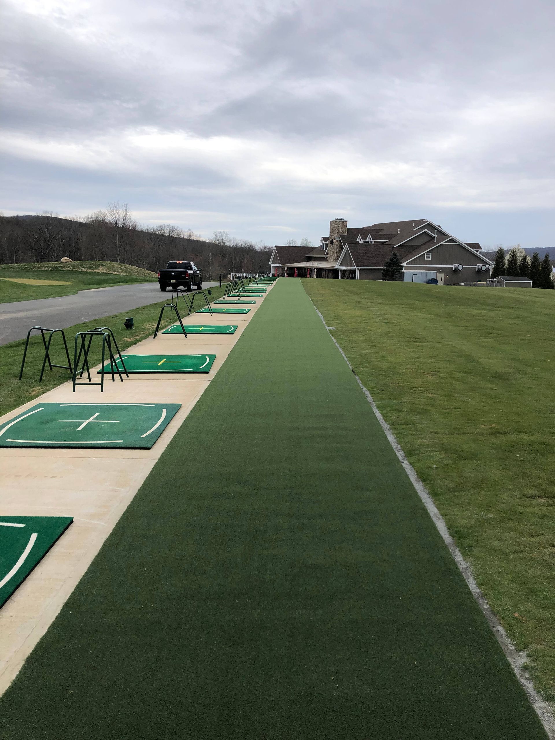 An outdoor driving range with lined-up hitting mats on a paved path, leading toward a distant clubhouse under a cloudy sky.