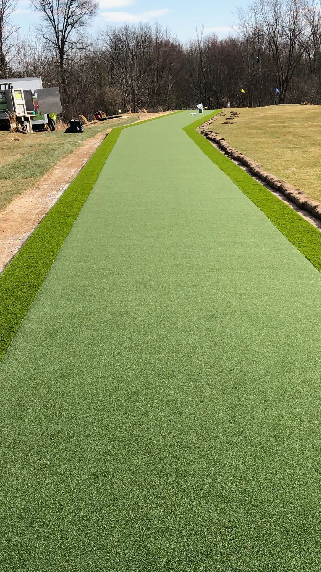 A long, straight path of bright green artificial turf framed by strips of natural grass, stretching toward distant trees.