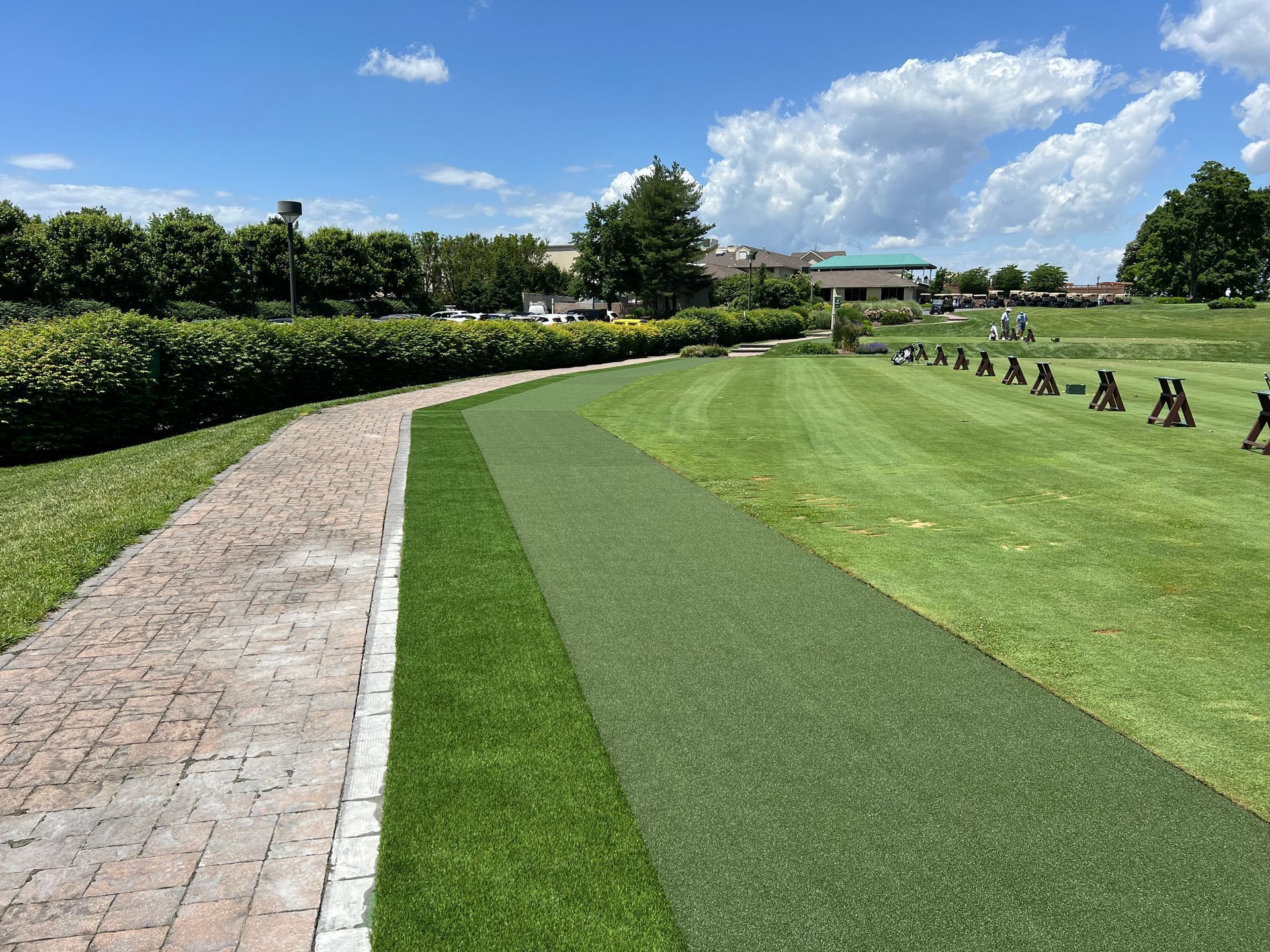 A brick pathway runs alongside an artificial turf strip and a grassy field with golf practice stands under a blue sky.