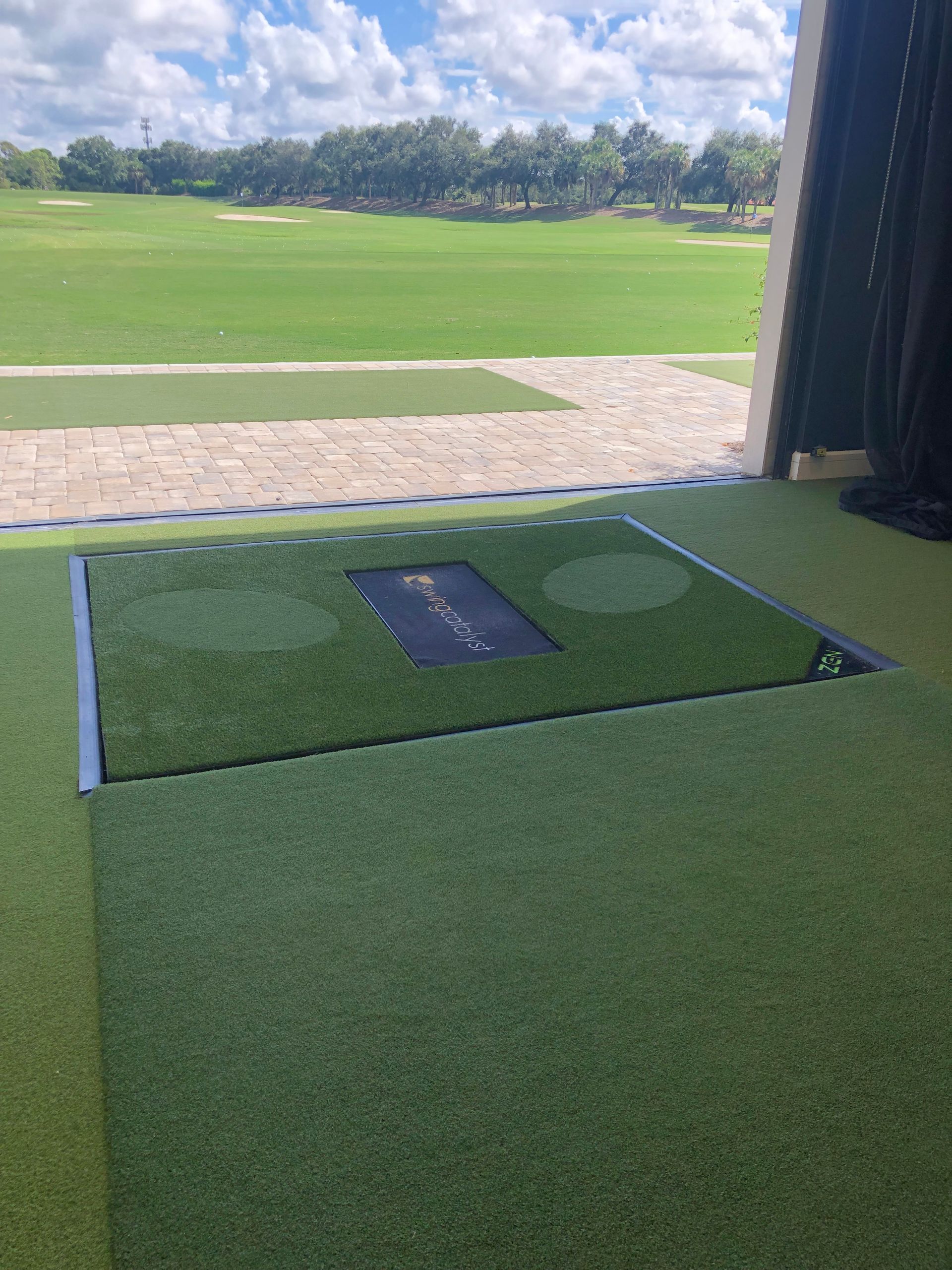 A golf hitting bay featuring an artificial turf mat and a golf ball dispenser, overlooking a green course under blue skies.