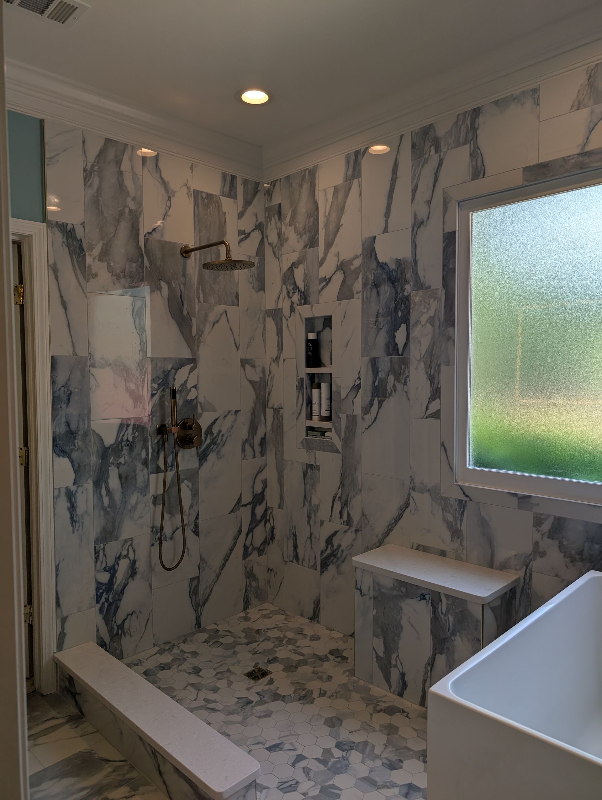 Shower with marble-like tile, built-in bench, and rain shower head. Features a window with a green outdoor view.