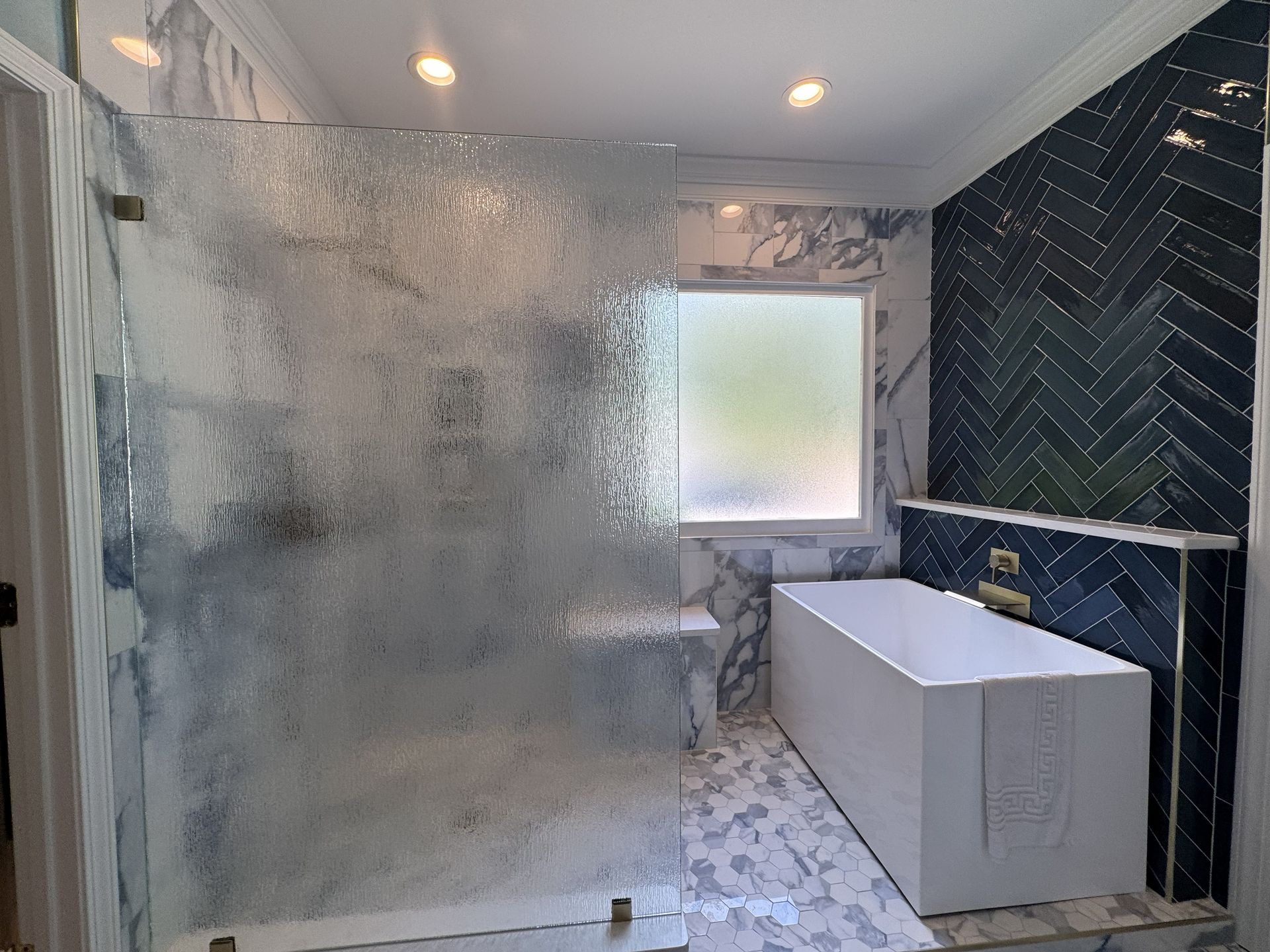 Modern bathroom with marble and blue tile, glass shower, white tub, and frosted window.
