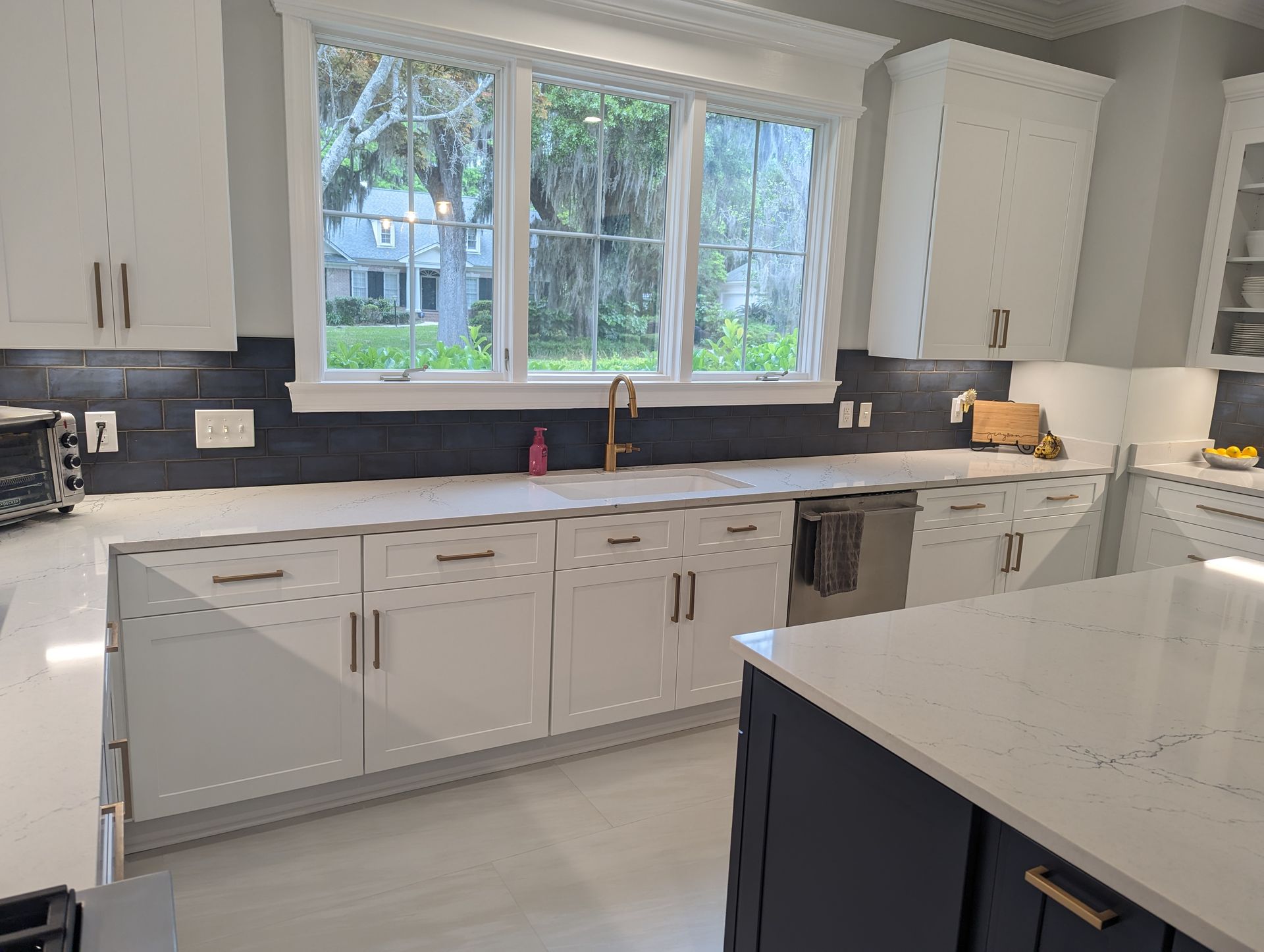 Modern white kitchen with island, quartz countertops, and a window overlooking a yard.