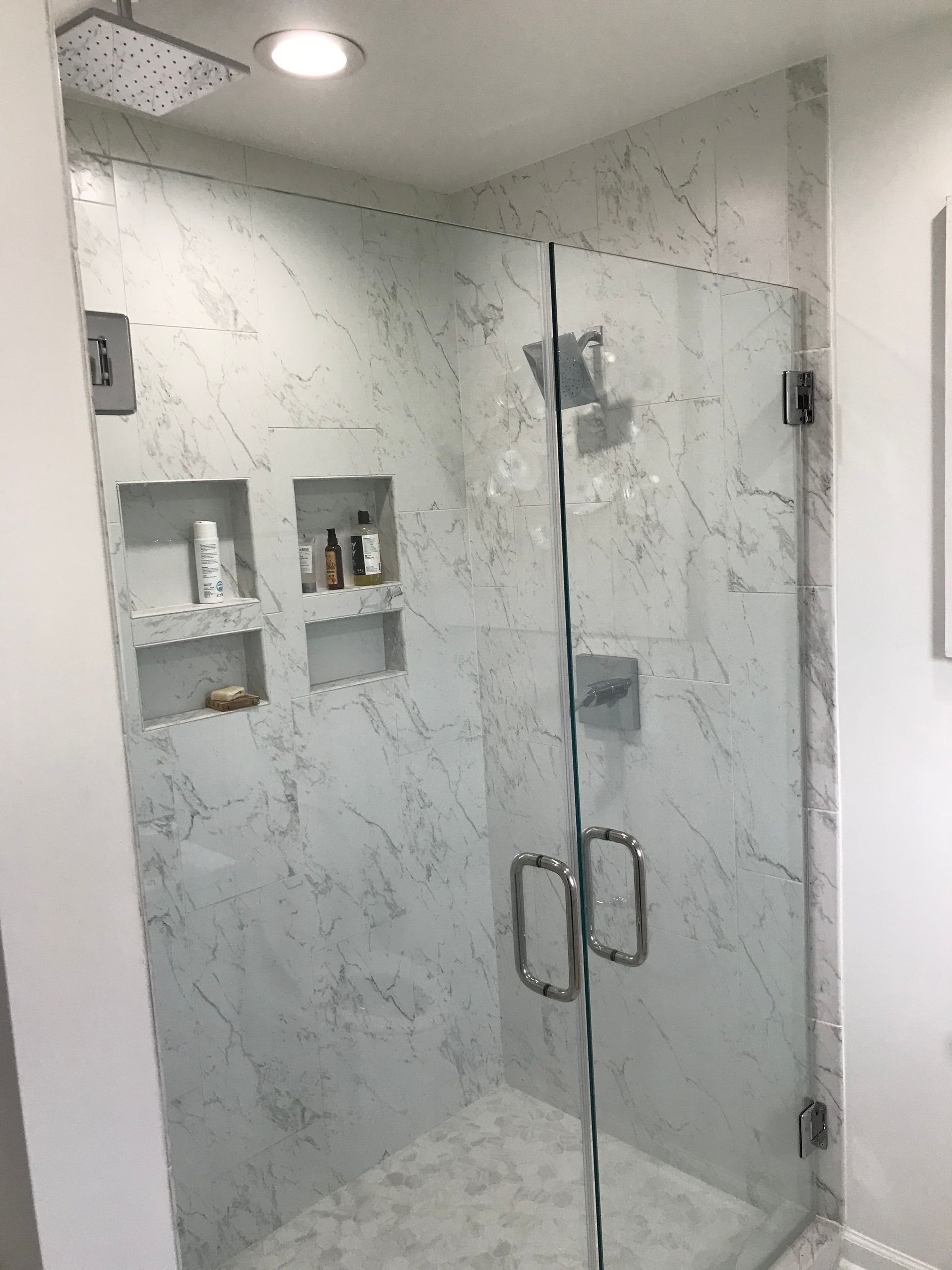 Shower with glass doors and white marble tile. Built-in shelves for toiletries.