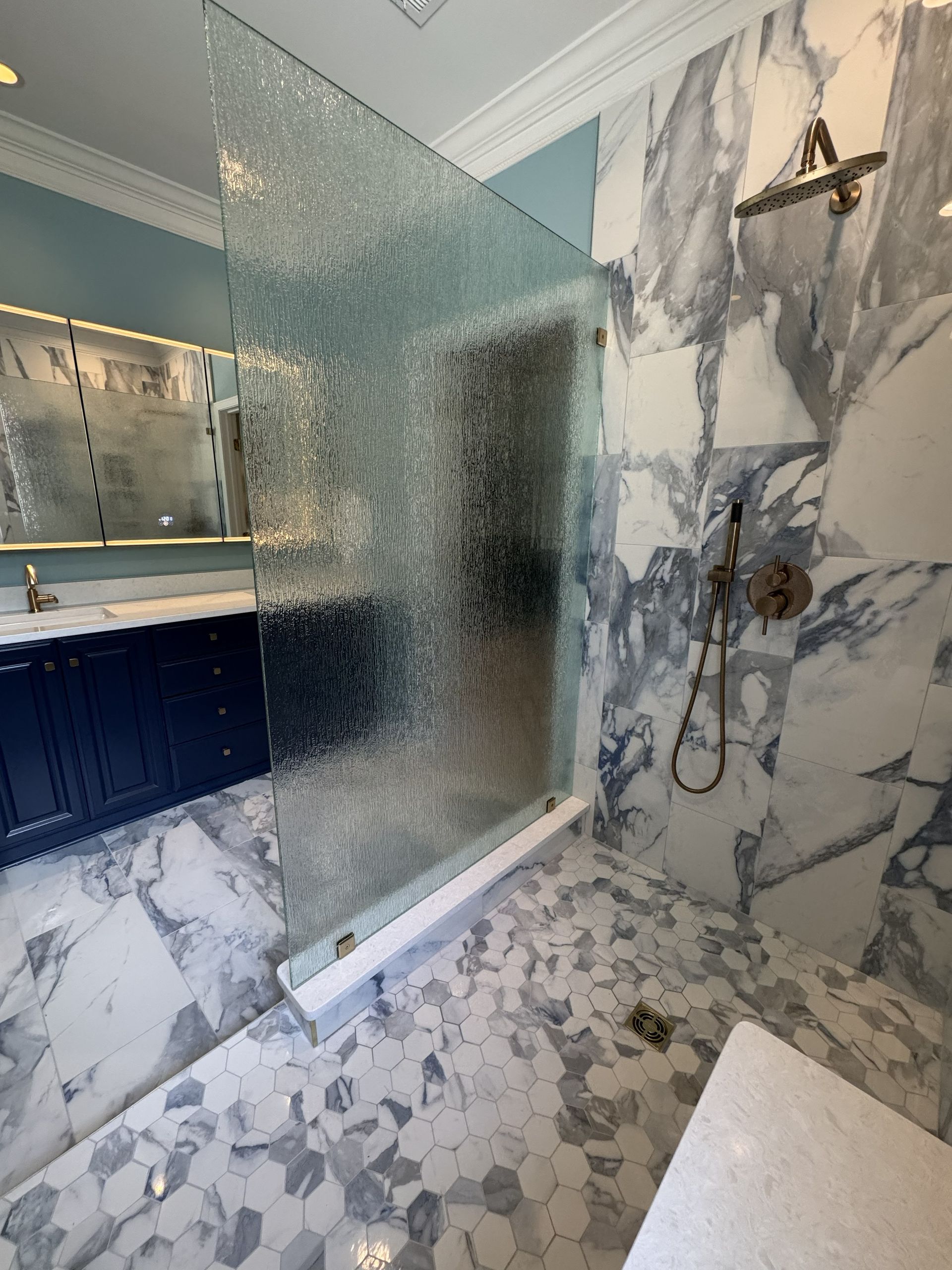 Shower with textured glass divider, marble walls and floor, blue vanity, and gold fixtures.