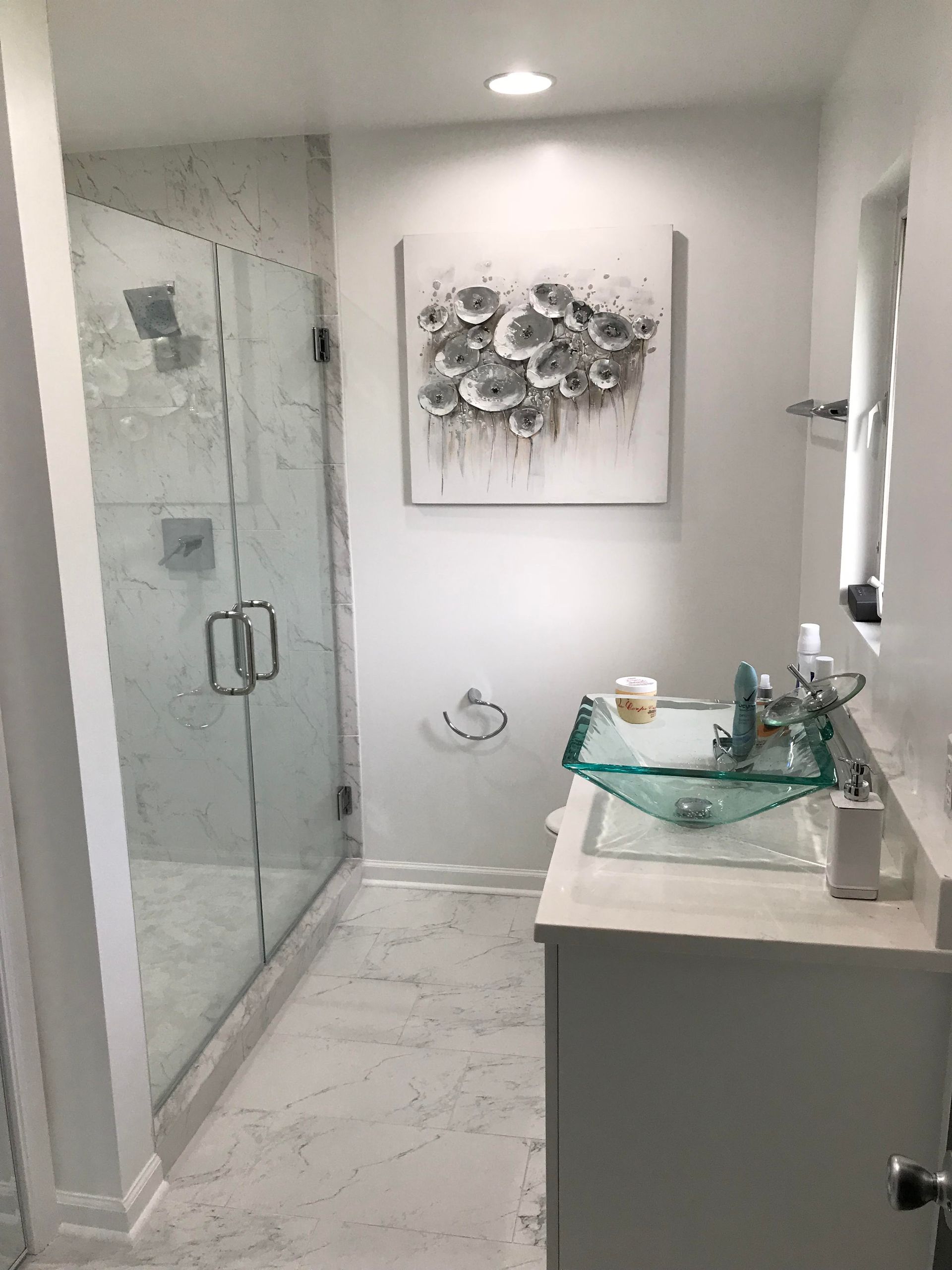 Bathroom with glass shower, white walls, marble floor, glass sink, and floral artwork.