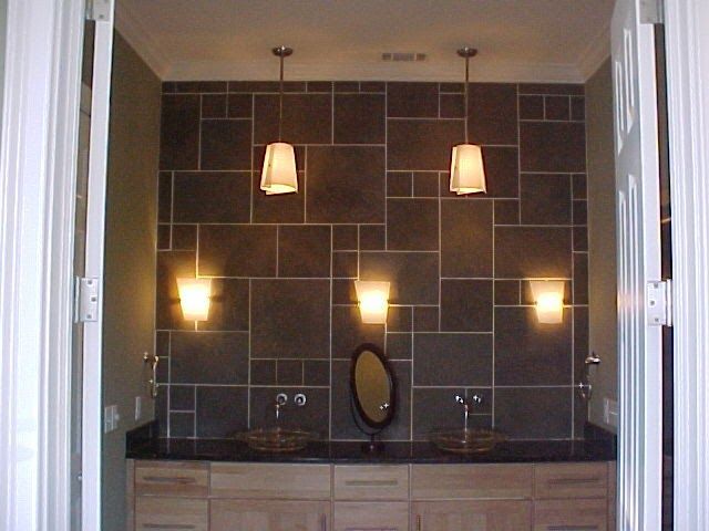 Bathroom with gray tile wall, pendant and wall sconce lighting, two sinks, oval mirror, and wooden vanity.