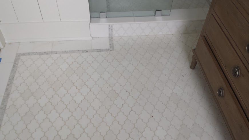 Bathroom floor with white and patterned tile, bordered by darker tile. A wooden dresser is in the corner.