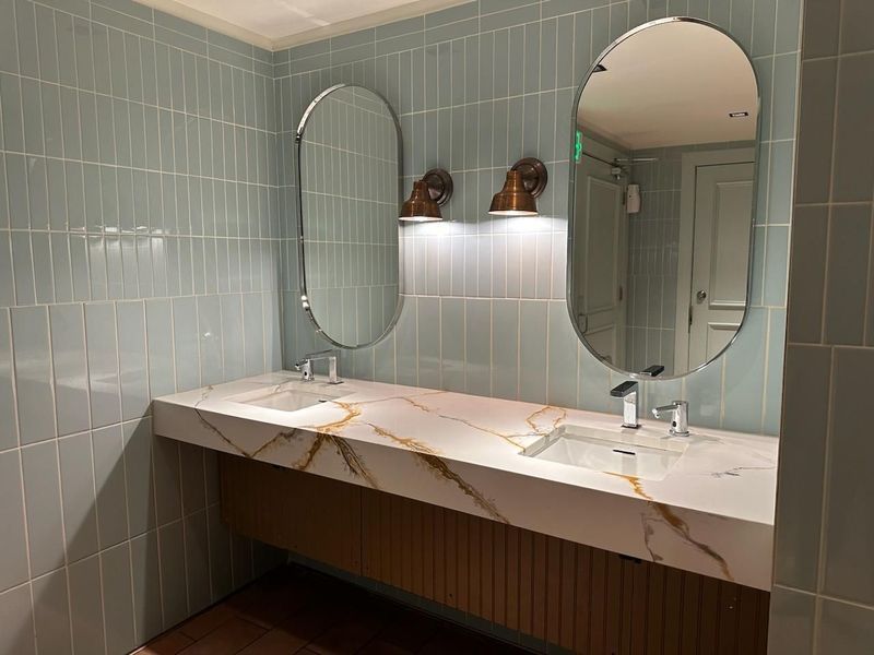 Bathroom with two sinks, oval mirrors, and light blue tiled walls. The countertop has gold veining.