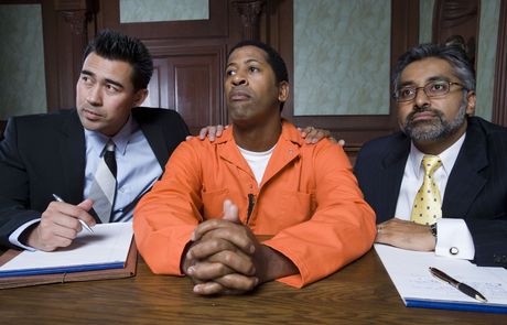 A man in an orange jumpsuit is sitting at a table with two men in suits.