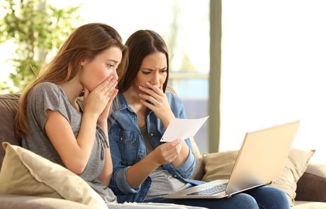 Two women are sitting on a couch looking at a laptop and a piece of paper.