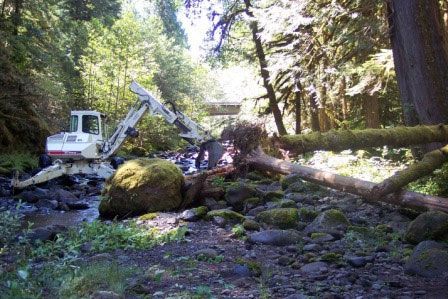 A white excavator is moving a large rock in the middle of a forest.