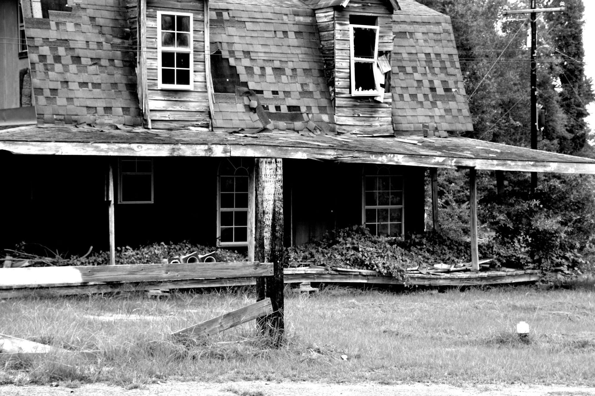 Black-and-white photo of a dilapidated house with a sagging roof, porch, and overgrown yard