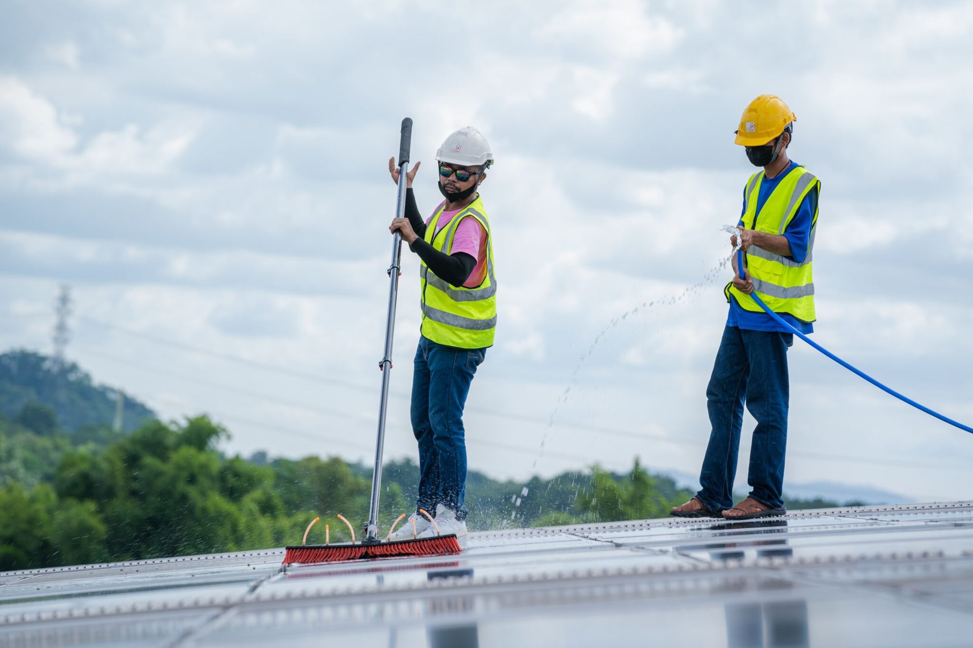 Two workers in hard hats and safety vests on a rooftop, using cleaning equipment against a cloudy sky