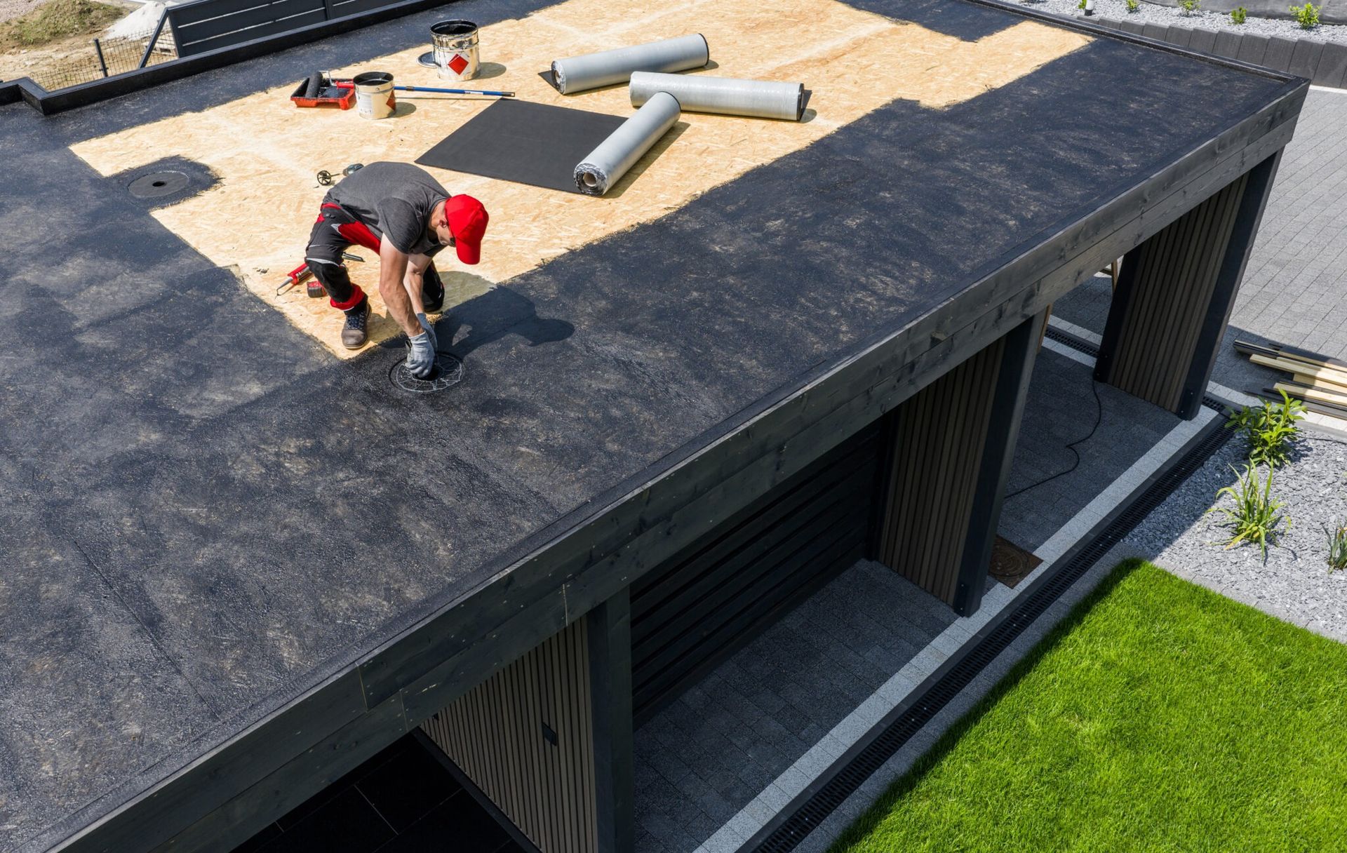 Person kneeling on a rooftop beside gray pipes and vents, with a green lawn below.