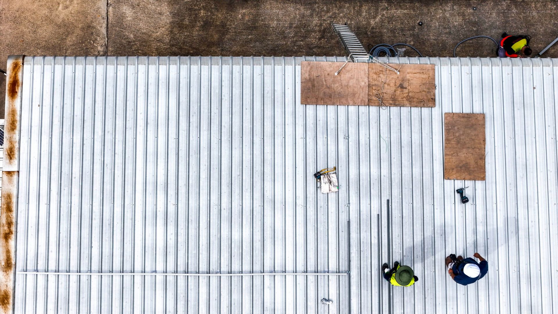 Overhead view of three workers on a white corrugated roof, carrying brown panels and tools.