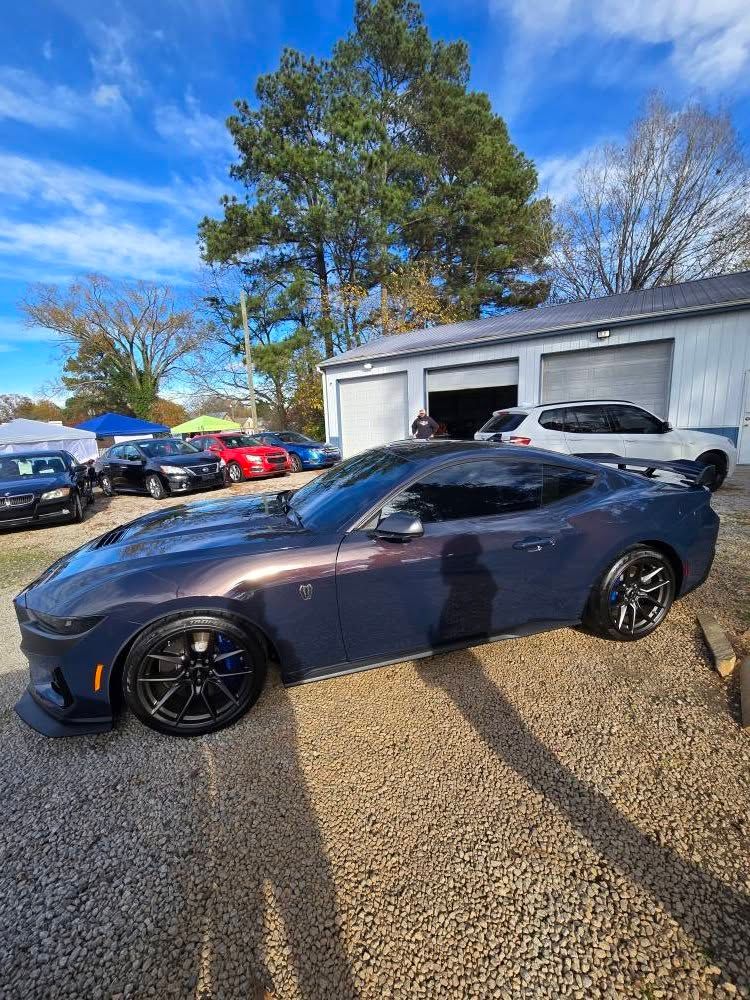 A gray sports car is parked in a gravel lot in front of a garage.