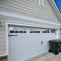 A white garage door is sitting on the side of a house next to a green trash can.