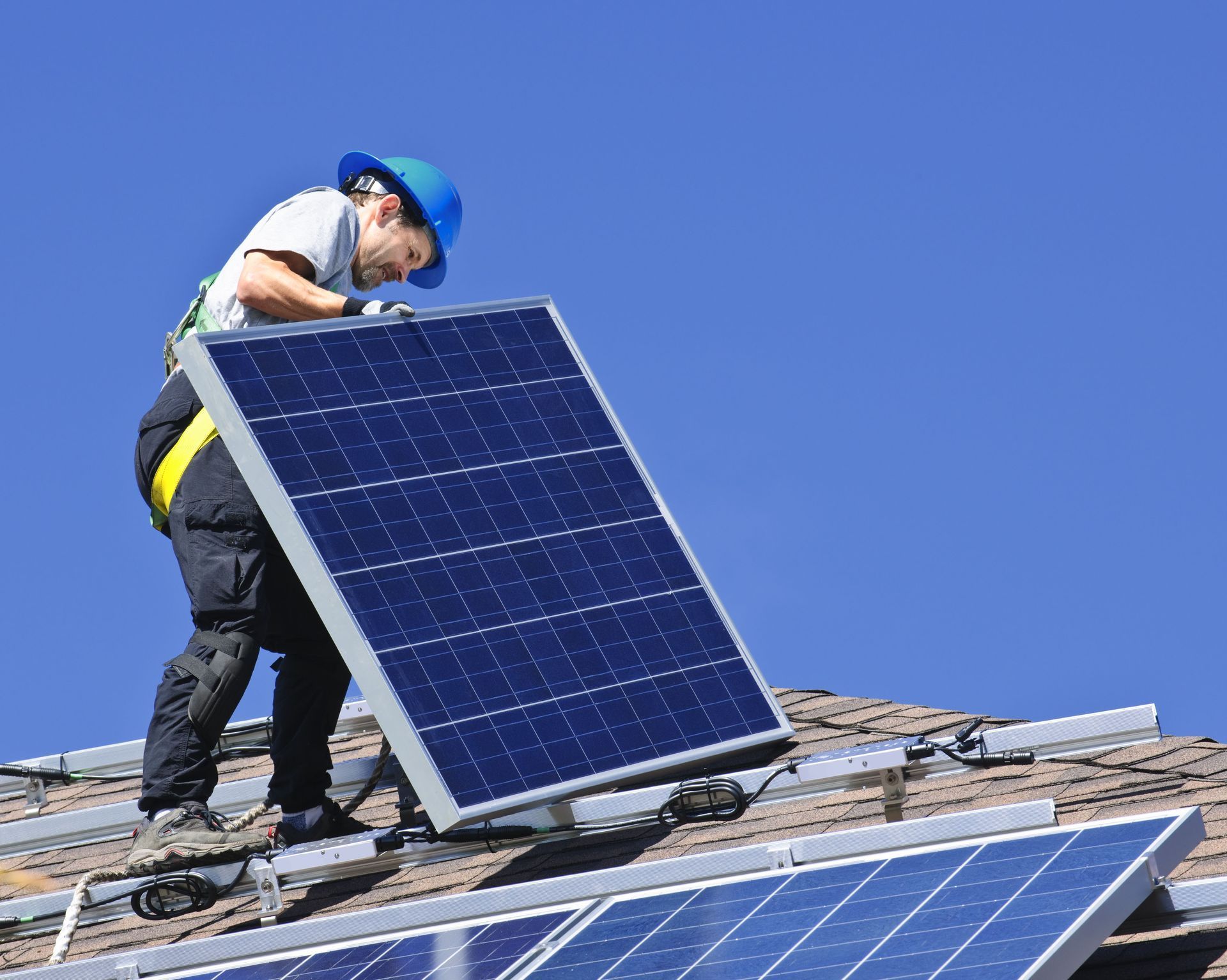Rooftop solar panel installation: Worker in blue hard hat securing a panel on a residential roof against a blue sky.