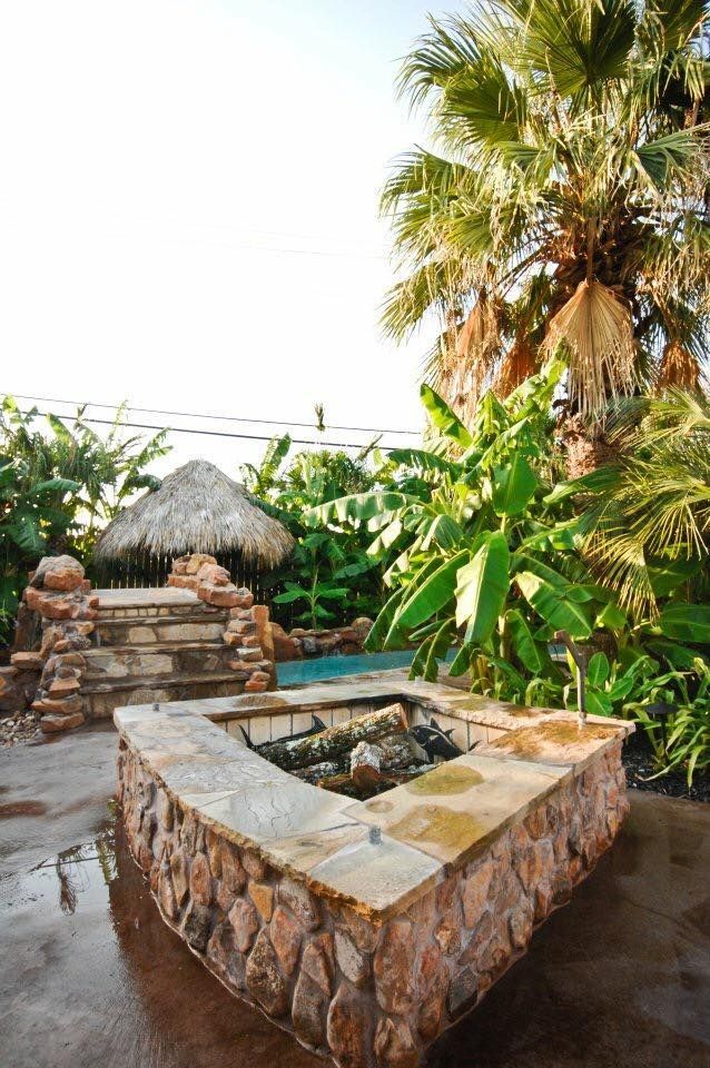 A stone fire pit is surrounded by palm trees and a thatched hut.