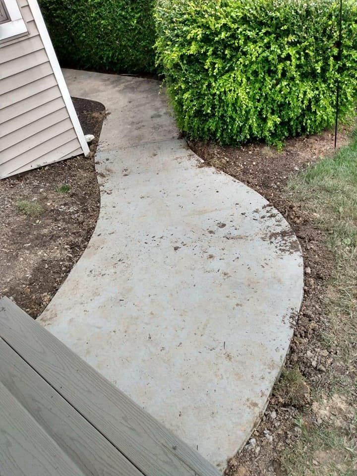 A concrete walkway leading to a house next to a hedge.