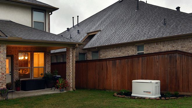 A beige standby generator on a concrete pad in a grassy yard, surrounded by greenery.