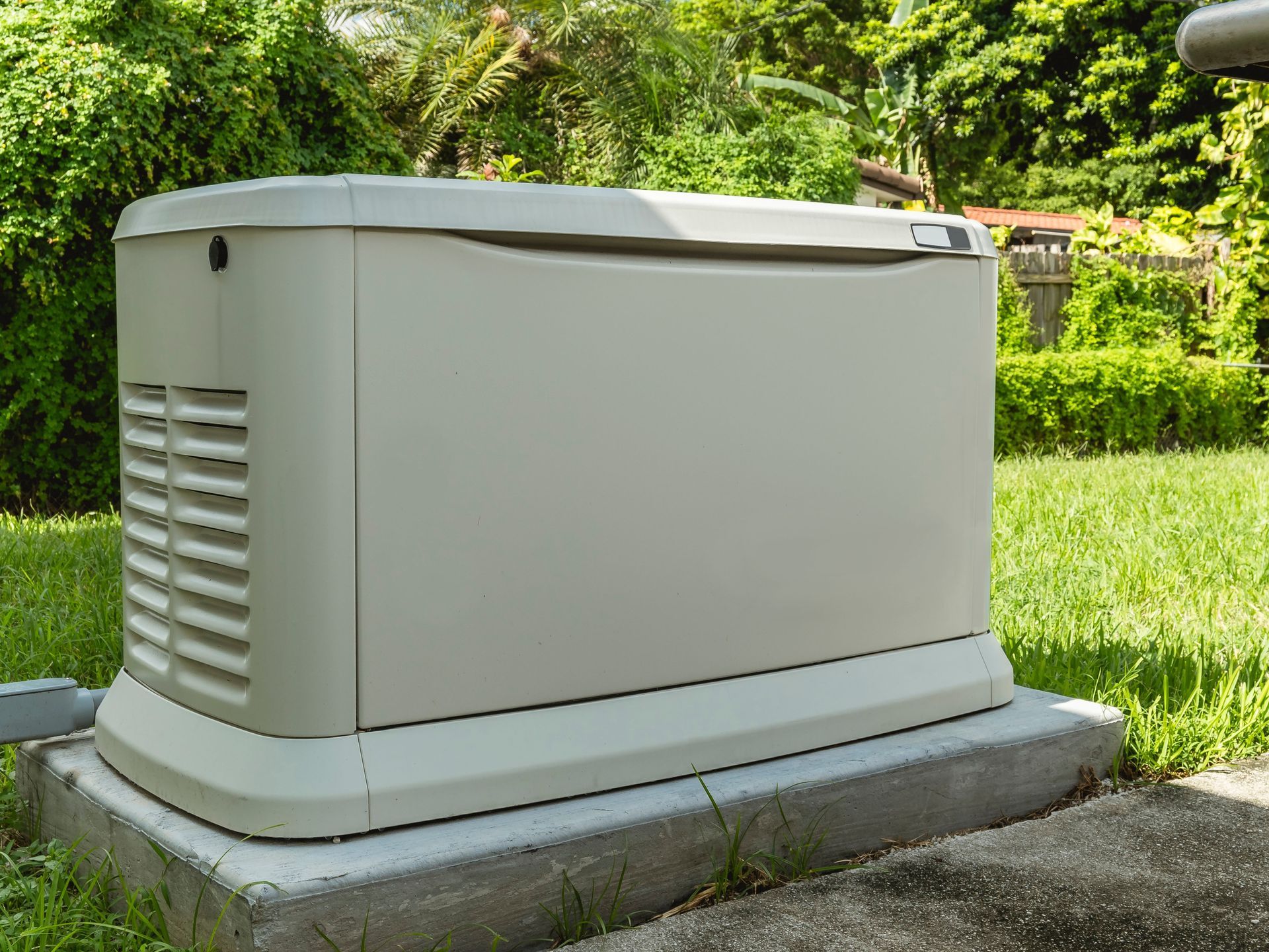 A beige standby generator on a concrete pad in a grassy yard, surrounded by greenery.