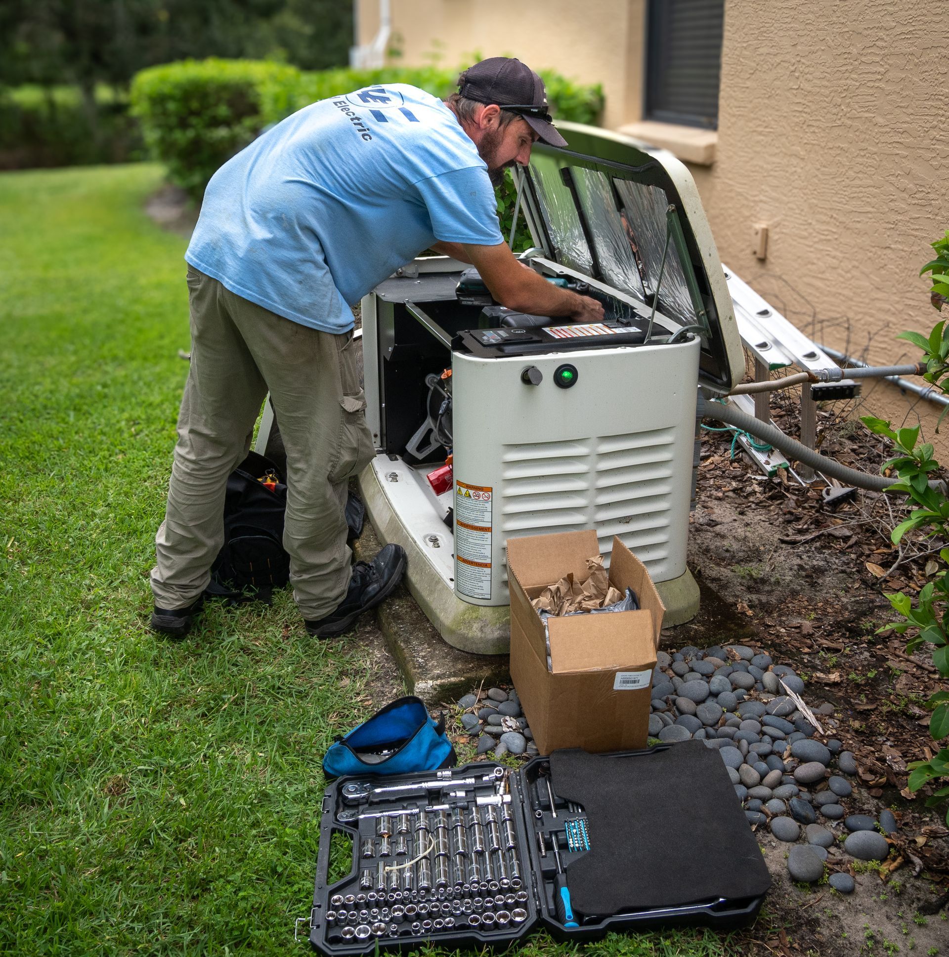 Technician in a light blue shirt works on an open outdoor residential backup generator next to a toolbox and box of parts.