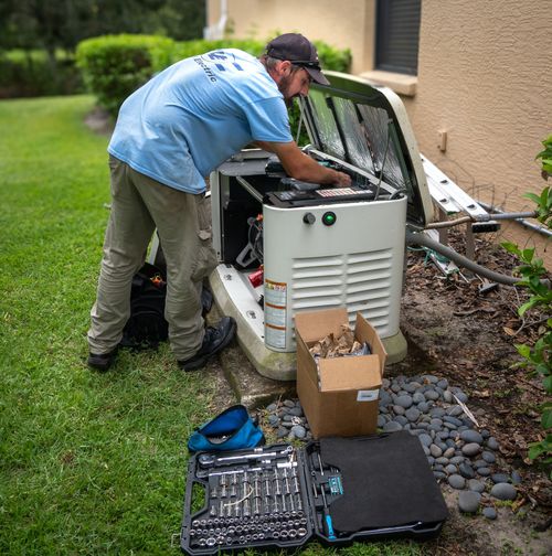 Technician in a light blue shirt works on an open outdoor residential backup generator next to a toolbox and box of parts.