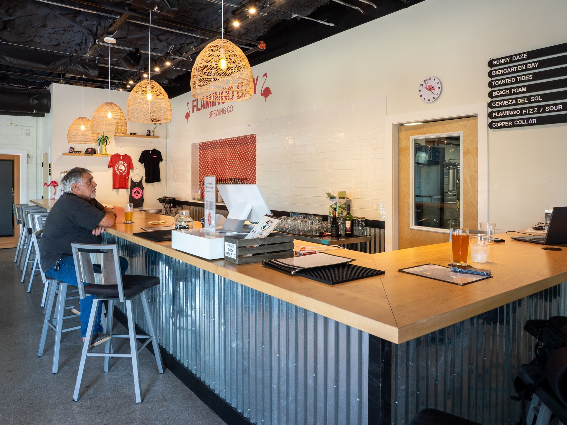 An indoor bar with a wood counter, corrugated metal base, and pendant lights, with a person sitting on a stool.