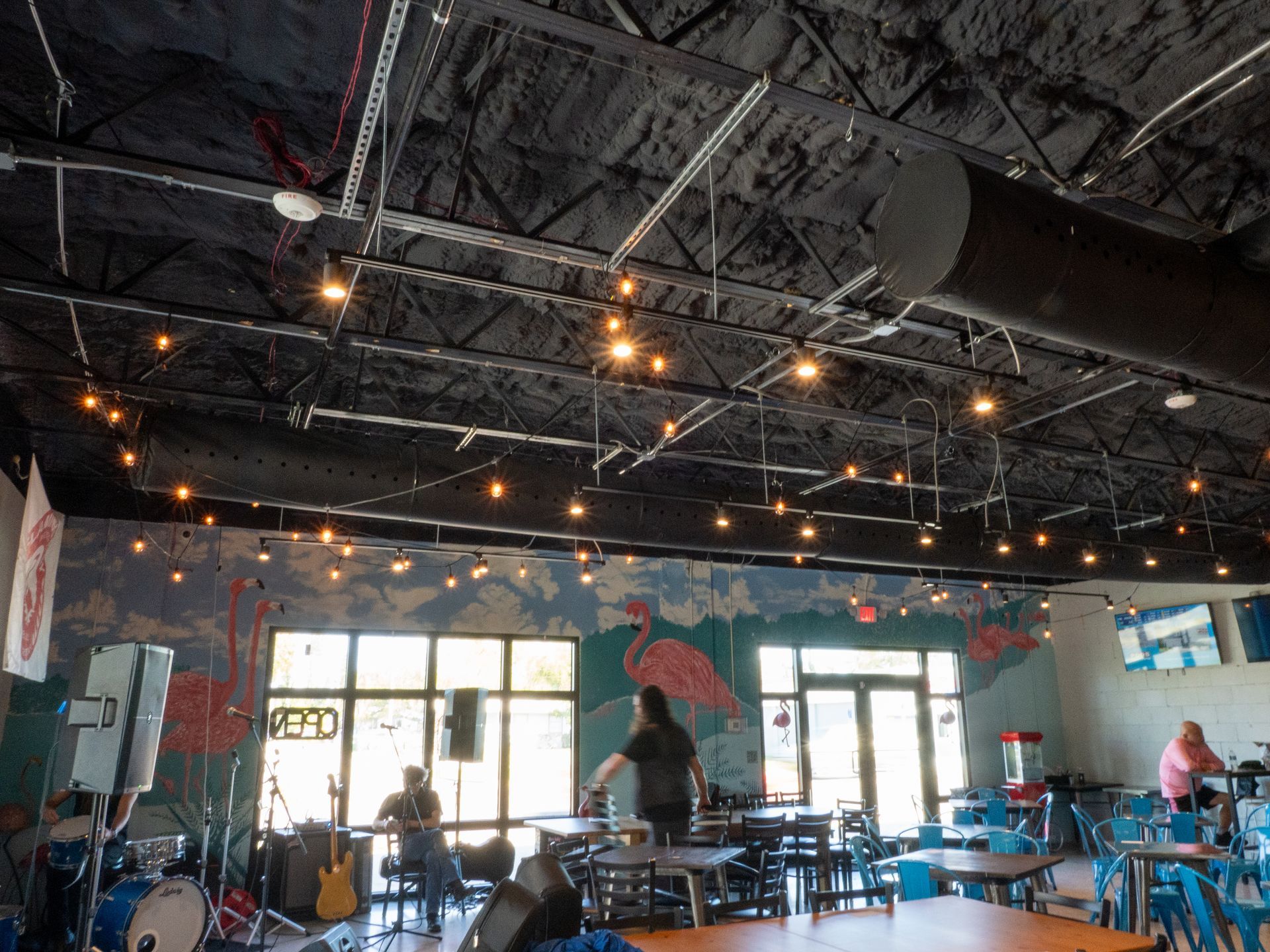 An interior view of a restaurant with high ceilings, string lights, and a mural featuring pink flamingos.